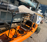 An orange sit-on-top kayak with a gray bimini and black seat is photographed outdoors beside a building. The kayak has mounted accessories and appears to be photographed in a real environment rather than a studio setup.