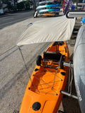 An orange sit-on-top kayak with a beige canopy and black seat is photographed outdoors beside a building. The kayak has mounted accessories and appears to be parked on a lift or rack in a real outdoor setting with other boats and buildings visible in the background.