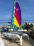 A white catamaran sailboat on a trailer outdoors, featuring a tall colorful sail with vertical text reading 'HOBIE' and a large geometric pattern in blue, green, yellow, red, and purple. The boat is photographed on a gravel lot with buildings, trees, and utility poles in the background under a blue sky.