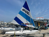 A white SOL catamaran sailboat on a trailer, featuring a blue and white striped mainsail with the SOL logo, photographed outdoors at a marina under a blue sky.