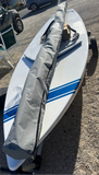 A white sailboard with blue stripes resting on a trailer outdoors, with a gray sail cover laid diagonally across the board. The setting is a gravel parking lot with a truck, beach chair, and cars in the background.