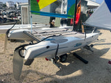 A white hobie wave catamaran sailboat on a trailer, photographed outdoors on a gravel surface with other boats and a colorful sail visible in the background.