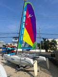 A Hobie Wave catamaran sailboat on a trailer outdoors under a blue sky, with a large colorful sail featuring vertical 'HOBIE' lettering and a stylized 'H' logo.